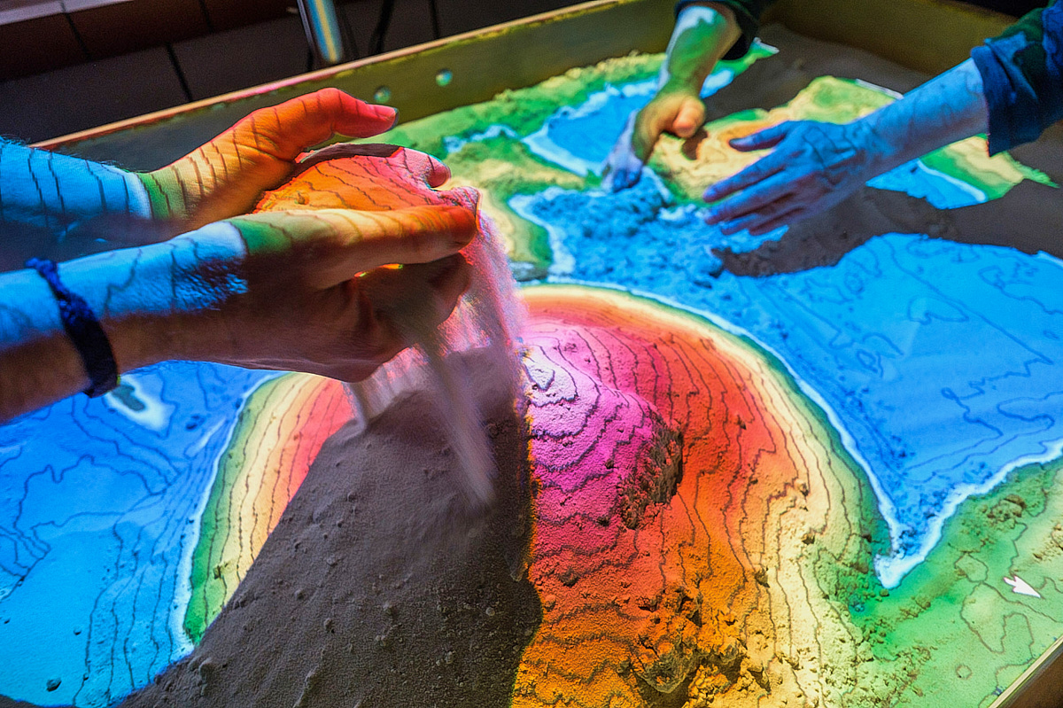 Zwei Wissenschaftler schichten Sand in einer Experiment aufeinander, um Höhenlinien sichtbar zu machen.