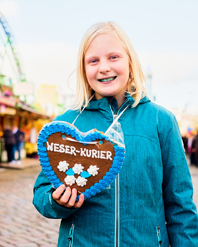 Die junge Gewinnerin eines Lesewettbewerbs der Kinderzeitung des Weser-Kuriers hält auf dem Freimarkt ein angebissenes Lebkuchenherz in die Kamera.