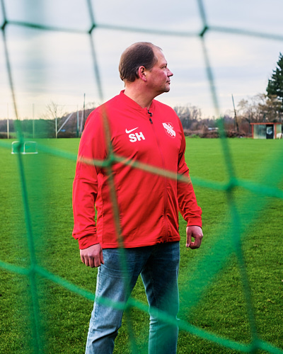 Pressefotografie von Fußballtrainer Sven Havekost, der hinter dem Netz eines Fußballtores cool in die Ferne blickt.