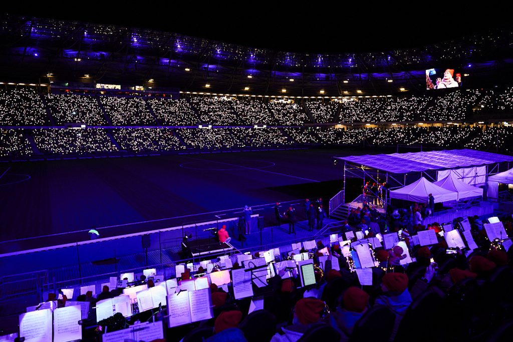 Die Eventfotografie zeigt ein volles Stadion am Abend während des Stadionsingens, während Menschen ihre Handylichter in die Höhe halten.